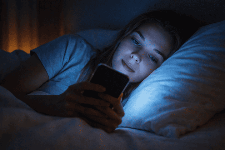 Personne utilisant un smartphone dans son lit la nuit éclairée par la lumière bleue de l’écran.
