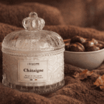 Bougie parfumée à la Châtaigne de L'Échoppe Buissonnière posée sur un plaid en laine avec des châtaignes et des feuilles d'automne
