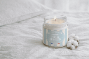 Bougie parfumée Fleur de Coton de L'Échoppe Buissonnière allumée sur un drap blanc avec des fleurs de coton