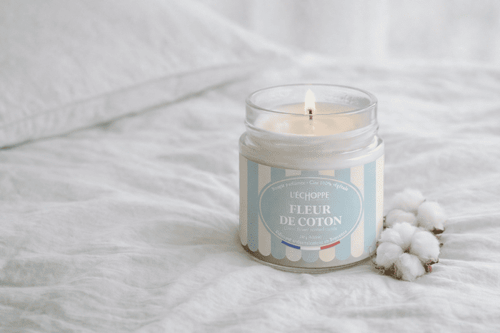 Bougie parfumée Fleur de Coton de L'Échoppe Buissonnière allumée sur un drap blanc avec des fleurs de coton