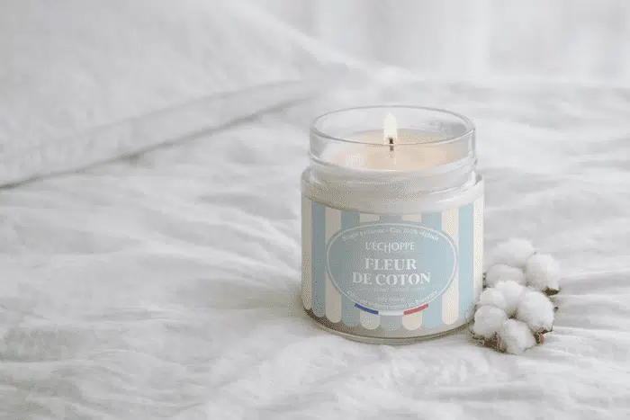 Bougie parfumée Fleur de Coton de L'Échoppe Buissonnière allumée sur un drap blanc avec des fleurs de coton