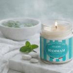 Bougie parfumée Hammam de L'Échoppe Buissonnière allumée sur un plan de marbre avec des feuilles de menthe et des galets blancs