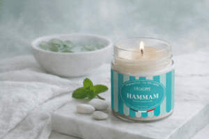 Bougie parfumée Hammam de L'Échoppe Buissonnière allumée sur un plan de marbre avec des feuilles de menthe et des galets blancs