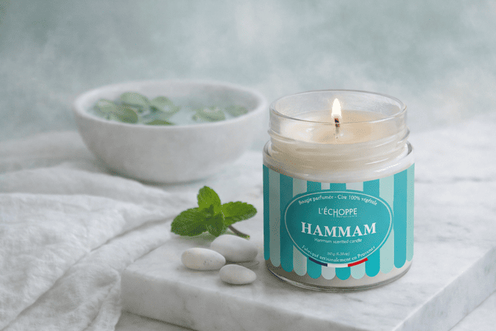Bougie parfumée Hammam de L'Échoppe Buissonnière allumée sur un plan de marbre avec des feuilles de menthe et des galets blancs