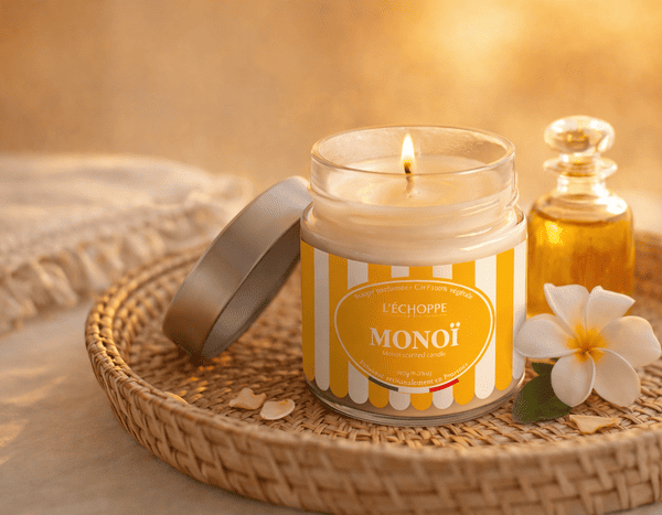 Bougie parfumée Monoï de L'Échoppe Buissonnière allumée sur un plateau en osier avec une fleur de tiaré et un flacon d'huile