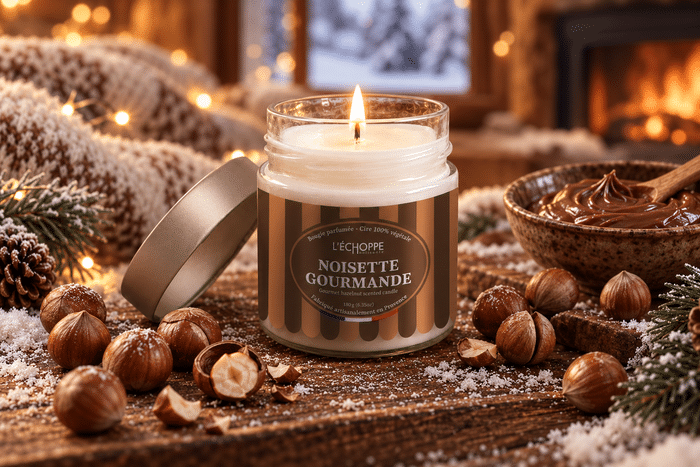 Bougie parfumée noisette gourmande échoppe buissonnière allumée Bougie parfumée Noisette Gourmande L'Échoppe Buissonnière allumée sur un plan en bois avec des noisettes entières et concassées