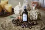 Brume végétale Chocolat Noisette Atelier Louis en flacon verre ambré avec vaporisateur noir, posée sur un set rond en fibres naturelles parmi chocolat aux noisettes, marrons et pomme de pin, avec des bougies et des citrouilles décoratives en arrière-plan