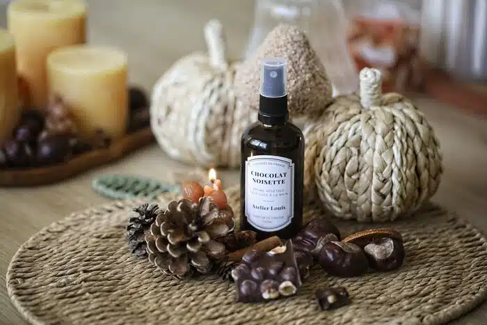 Brume végétale Chocolat Noisette Atelier Louis en flacon verre ambré avec vaporisateur noir, posée sur un set rond en fibres naturelles parmi chocolat aux noisettes, marrons et pomme de pin, avec des bougies et des citrouilles décoratives en arrière-plan