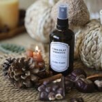 Flacon ambré de brume d'ambiance Chocolat Noisette Atelier Louis 100 ml, entouré d'une pomme de pin, d'un carré de chocolat aux noisettes, de marrons et d'une petite bougie allumée, devant des citrouilles en fibres tressées
