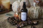 Flacon ambré de brume d'ambiance Chocolat Noisette Atelier Louis 100 ml, entouré d'une pomme de pin, d'un carré de chocolat aux noisettes, de marrons et d'une petite bougie allumée, devant des citrouilles en fibres tressées