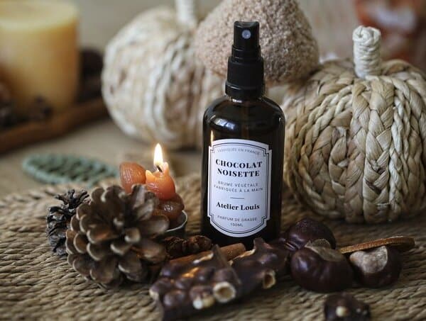 Flacon ambré de brume d'ambiance Chocolat Noisette Atelier Louis 100 ml, entouré d'une pomme de pin, d'un carré de chocolat aux noisettes, de marrons et d'une petite bougie allumée, devant des citrouilles en fibres tressées