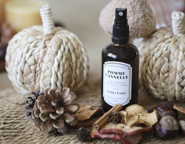 Flacon ambré de brume d'ambiance Pomme Cannelle Atelier Louis 100 ml, posé sur un set en fibres tressées entre deux citrouilles décoratives, avec pommes de pin, bâtons de cannelle, rondelles de pomme séchée et marrons