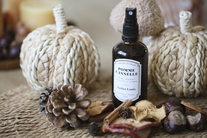 Brume Pomme Cannelle Atelier Louis, décor d'automne Flacon ambré de brume d'ambiance Pomme Cannelle Atelier Louis 100 ml, posé sur un set en fibres tressées entre deux citrouilles décoratives, avec pommes de pin, bâtons de cannelle, rondelles de pomme séchée et marrons